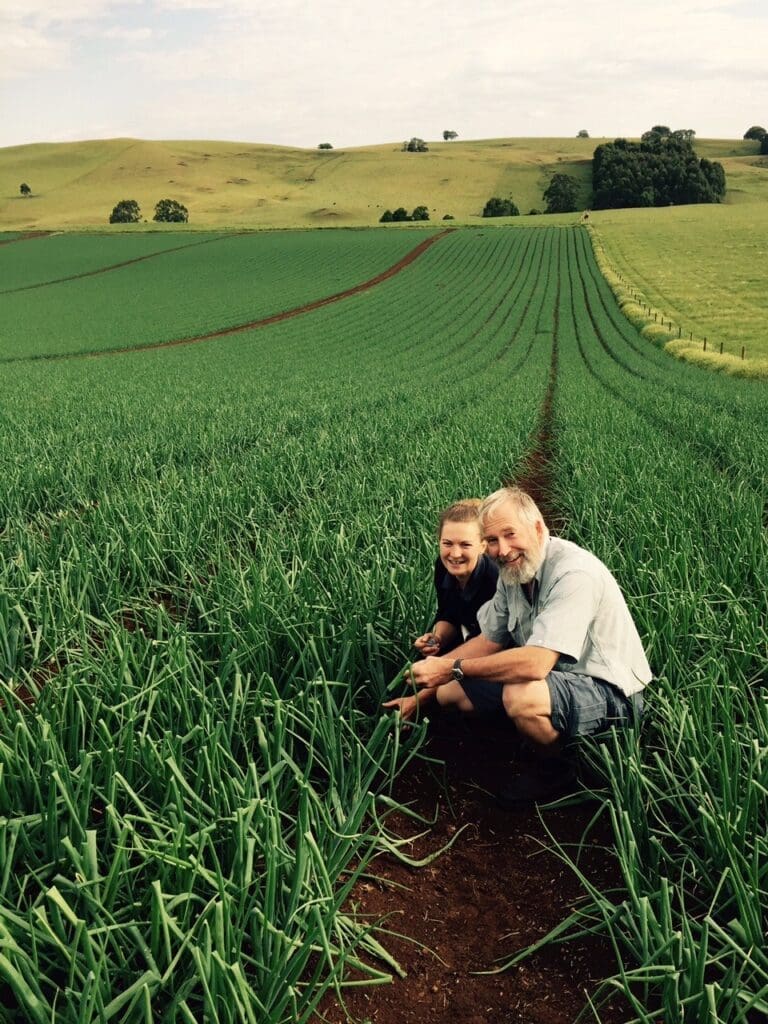 Two people crouching in a green field with hills in the background, examining crops.